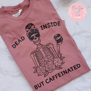 May include: A pink t-shirt with a black graphic of a skeleton holding a coffee cup. The text "Dead Inside But Caffeinated" is printed on the shirt.