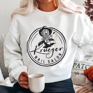 Puede incluir: Sudadera blanca con un dise&ntilde;o gr&aacute;fico en blanco y negro de un personaje de pel&iacute;cula de terror sosteniendo herramientas de manicura, con las palabras "Krueger Nail Salon" en un dise&ntilde;o circular.