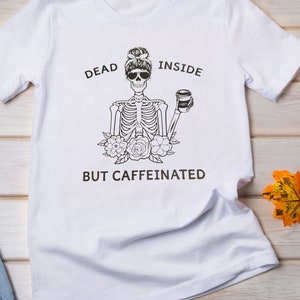 May include: White t-shirt with a black and white skeleton graphic holding a coffee cup. The text "Dead Inside But Caffeinated" is printed on the shirt.