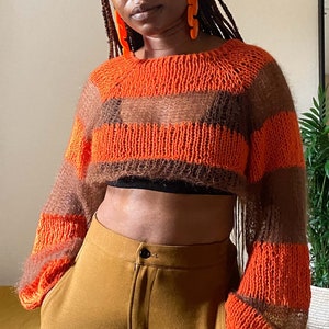 May include: A cropped, striped sweater in shades of orange and brown. The sweater has long sleeves and a round neckline. The model is wearing brown trousers. The sweater is a handmade, knitted item.