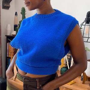 May include: A bright blue sleeveless knitted sweater with a round neckline. The sweater is cropped and has a loose fit.