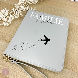 Könnte beinhalten: Graue Reißverschlusstasche für Reisen mit einer weißen Aufschrift "Familie Kallus" und einem schwarzen Flugzeug mit einem gepunkteten Linienzug, der eine Herzform bildet.