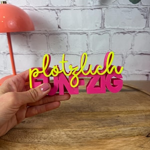 Könnte beinhalten: Ein dreidimensionales Schild mit dem Wort "plötzlich" in leuchtend gelber Schreibschrift, das über dem in Blockbuchstaben geschriebenen Wort "FÜNFZIG" in kräftigem Pink liegt. Das Schild wird in einer Hand gehalten und zeigt die Kontraste.