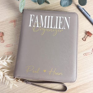 Könnte beinhalten: Ein grauer Familienplaner mit dem Text "FAMILIEN Organizer" in Weiß und Gold. Der Organizer hat eine Handschlaufe und die Namen "Paul" und "Henri" in Gold. Der Organizer liegt auf einer Holzoberfläche.