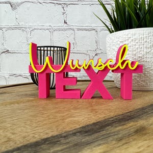 Könnte beinhalten: Ein leuchtend pinkfarbenes "TEXT"-Schild mit dem Wort "Wunsch" in gelber Schrift. Das Schild steht auf einer Holzoberfläche, im Hintergrund eine weiße Backsteinmauer und eine Topfpflanze.