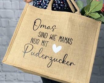 May include: A beige burlap tote bag with black script that reads "Omas sind wie Mamas nur mit Puderzucker" and a white heart. The bag has two handles and is being held by a person.