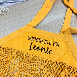 Könnte beinhalten: Senfgelbe Strandtasche mit Netzdesign. Die Tasche hat ein rechteckiges Stoffpaneel mit dem Text "SANDSPIELZEUG VON Leonie" in Schwarz. Die Tasche hat zwei Schultergurte.