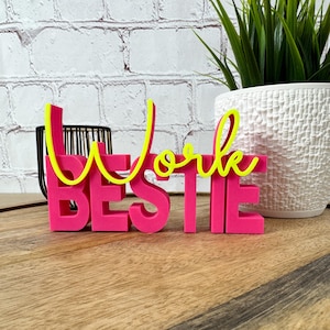 Könnte beinhalten: Ein dreidimensionales Schreibtischschild mit dem Wort BESTIE in kräftigen, leuchtend pinken Blockbuchstaben, überlagert von dem Wort Work in einer leuchtend gelben Schreibschrift, was ein farbenfrohes und auffälliges Dekorationsobjekt ergibt.