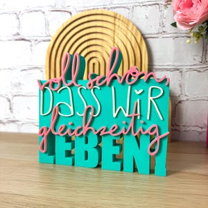 Könnte beinhalten: Ein türkisfarbenes Holzschild mit dem deutschen Satz "voll schön, dass wir gleichzeitig leben!" in rosa und weißen Buchstaben. Im Hintergrund befindet sich eine Holzdekoration in Regenbogenform.