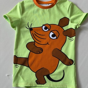 Könnte beinhalten: Ein lindgrünes Kurzarm-T-Shirt mit orangefarbenem Rundhalsausschnitt. Auf der Vorderseite befindet sich eine große Cartoon-Grafik einer braunen Maus mit dunkelbraunen Ohren, Füßen und Händen, die in einer dynamischen Pose dargestellt ist.
