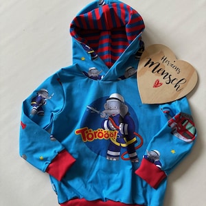 Könnte beinhalten: Blauer Kapuzenpullover mit roten Bündchen und rot-blau gestreiftem Kapuzenfutter. Auf der Vorderseite befindet sich ein Cartoon-Feuerwehrelfant mit dem Wort "Törööö!" und einer Feuerwehrwagen-Grafik. Ein Holzherz mit dem Text "Herzins Mensch" ist ebenfalls vorhanden.