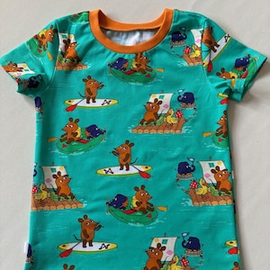 Könnte beinhalten: Ein kurzärmeliges T-Shirt in hellem Grün mit einem durchgehenden Muster aus braunen Mäusen auf Flößen, Stand-Up-Paddleboards und in kleinen Booten. Das T-Shirt hat einen einfarbigen orangefarbenen Rundhalsausschnitt und Cartoon-Motive.