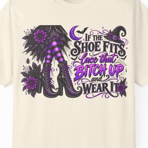 Op de afbeelding: Een crèmekleurig t-shirt met een grafische print van paars en zwart gestreepte kousen, zwarte veterschoenen, bloemen, een heksenhoed en een maansikkel. De tekst luidt: If the SHOE FITS lace that BITCH UP and WEAR IT.
