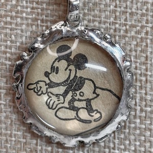 May include: A silver-colored pendant necklace featuring a black and white image of Mickey Mouse. The pendant has a textured edge and hangs from a black cord necklace.