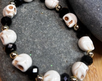 Pode incluir: Uma pulseira de contas com contas de caveira brancas, contas facetadas pretas e contas espaçadoras douradas. A pulseira é exibida em uma pedra cinza sobre um fundo de madeira. A pulseira é um acessório gótico ou com tema de Halloween.