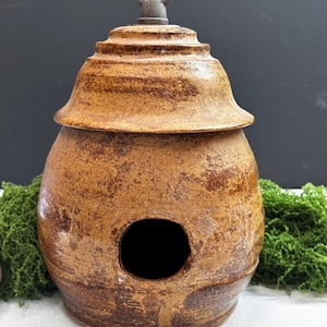 Pode incluir: Uma casa de pássaros de cerâmica redonda, castanha e texturizada com um acabamento manchado. Apresenta uma tampa larga e escalonada rematada com um pequeno anel de metal para pendurar. O corpo principal tem uma abertura circular central.