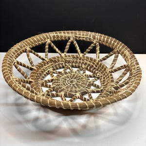 Pode incluir: Uma cesta redonda artesanal com um desenho de estrela no centro. A cesta &eacute; feita de fibras tecidas de cor natural, com um esquema de cores castanho claro e creme. O design vazado apresenta uma s&eacute;rie de raios radiantes.