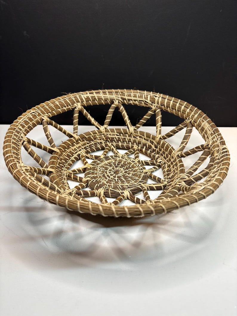 Pode incluir: Uma cesta redonda artesanal com um desenho de estrela no centro. A cesta &eacute; feita de fibras tecidas de cor natural, com um esquema de cores castanho claro e creme. O design vazado apresenta uma s&eacute;rie de raios radiantes.