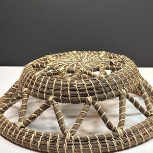 Pode incluir: Um cesto redondo artesanal com um design tecido. O cesto apresenta um esquema de cores castanho e bege, com um padr&atilde;o circular no topo e suportes triangulares nas laterais. Os materiais naturais criam uma est&eacute;tica r&uacute;stica.