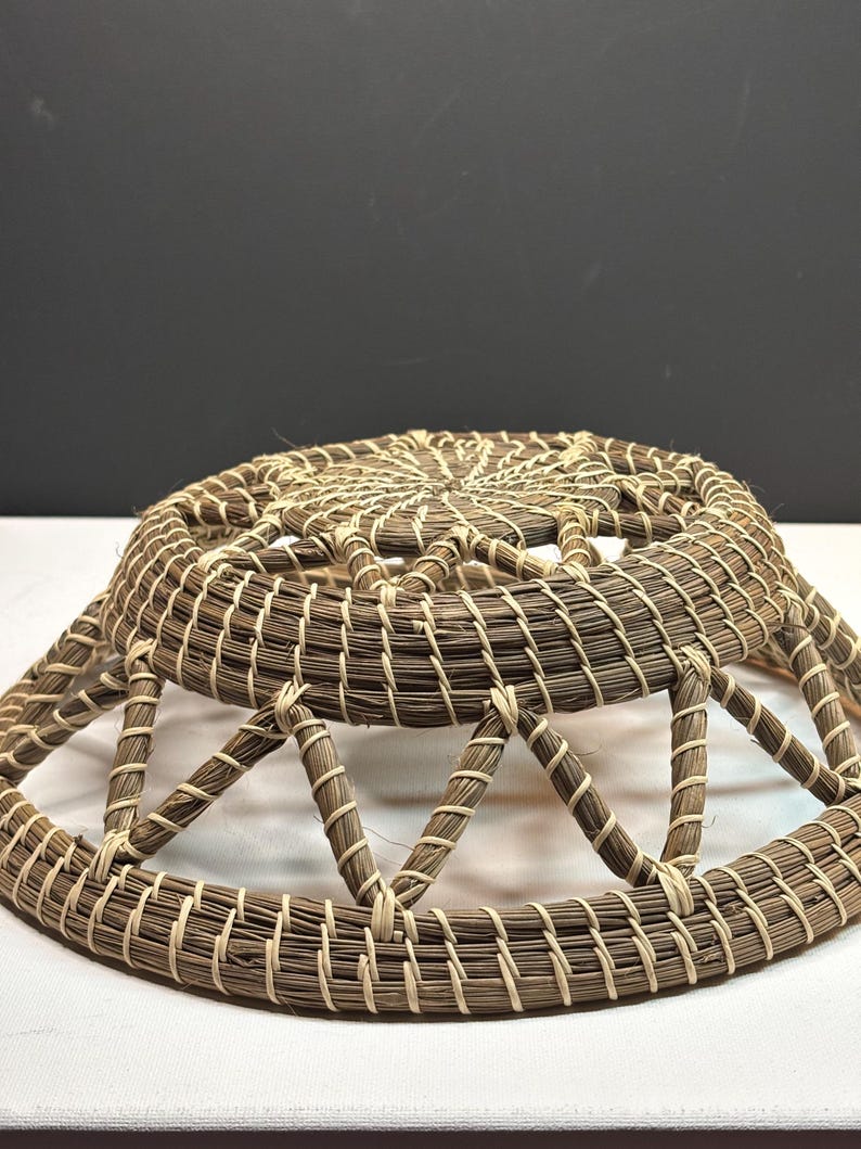 Pode incluir: Um cesto redondo artesanal com um design tecido. O cesto apresenta um esquema de cores castanho e bege, com um padr&atilde;o circular no topo e suportes triangulares nas laterais. Os materiais naturais criam uma est&eacute;tica r&uacute;stica.