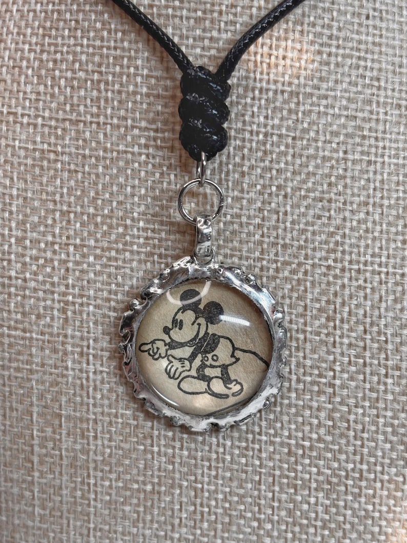 May include: A silver-colored pendant necklace featuring a black and white image of Mickey Mouse. The pendant has a textured edge and hangs from a black cord necklace.