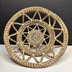 Pode incluir: Uma cesta redonda tran&ccedil;ada com um centro em espiral e um padr&atilde;o de treli&ccedil;a aberto em forma de estrela ao longo da borda externa. A cesta &eacute; feita de fibras naturais marrons e bege, exibindo uma textura artesanal detalhada.