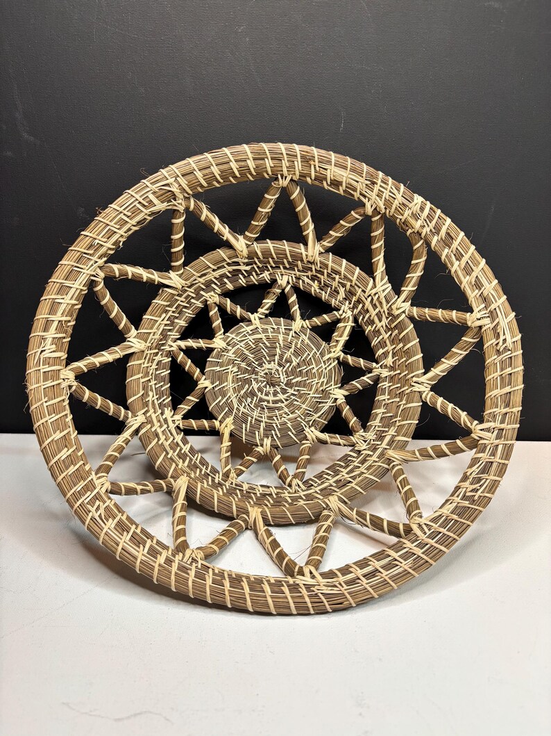 Pode incluir: Uma cesta redonda tran&ccedil;ada com um centro em espiral e um padr&atilde;o de treli&ccedil;a aberto em forma de estrela ao longo da borda externa. A cesta &eacute; feita de fibras naturais marrons e bege, exibindo uma textura artesanal detalhada.