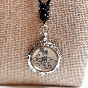 May include: A silver-toned pendant necklace featuring a circular charm with a vintage-style image of Mickey Mouse and Pluto. The pendant hangs from a black cord with an adjustable knot.