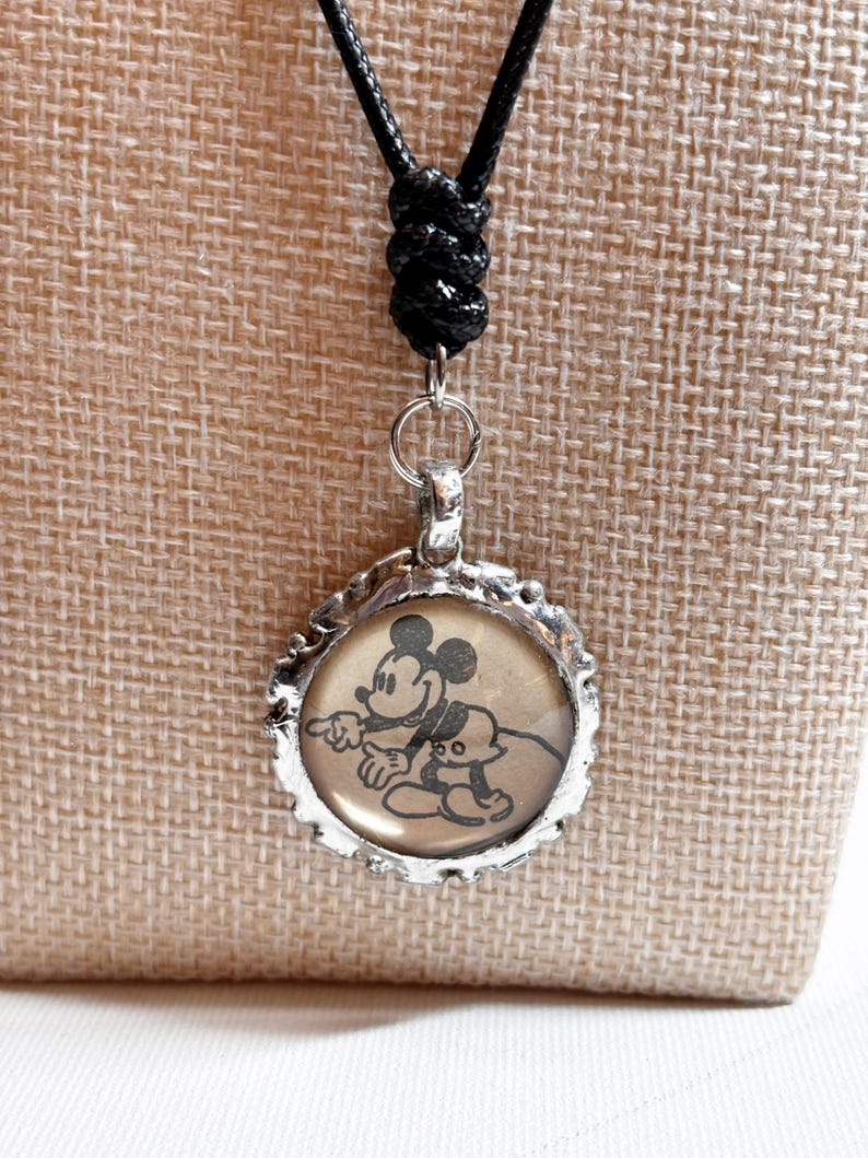 May include: A silver-toned pendant necklace featuring a circular charm with a vintage-style image of Mickey Mouse and Pluto. The pendant hangs from a black cord with an adjustable knot.