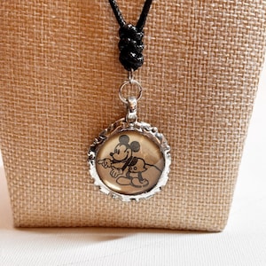 May include: A silver-toned pendant necklace featuring a circular charm with a black and white image of Mickey Mouse and Pluto. The charm is set in a decorative frame and hangs from a black cord necklace.