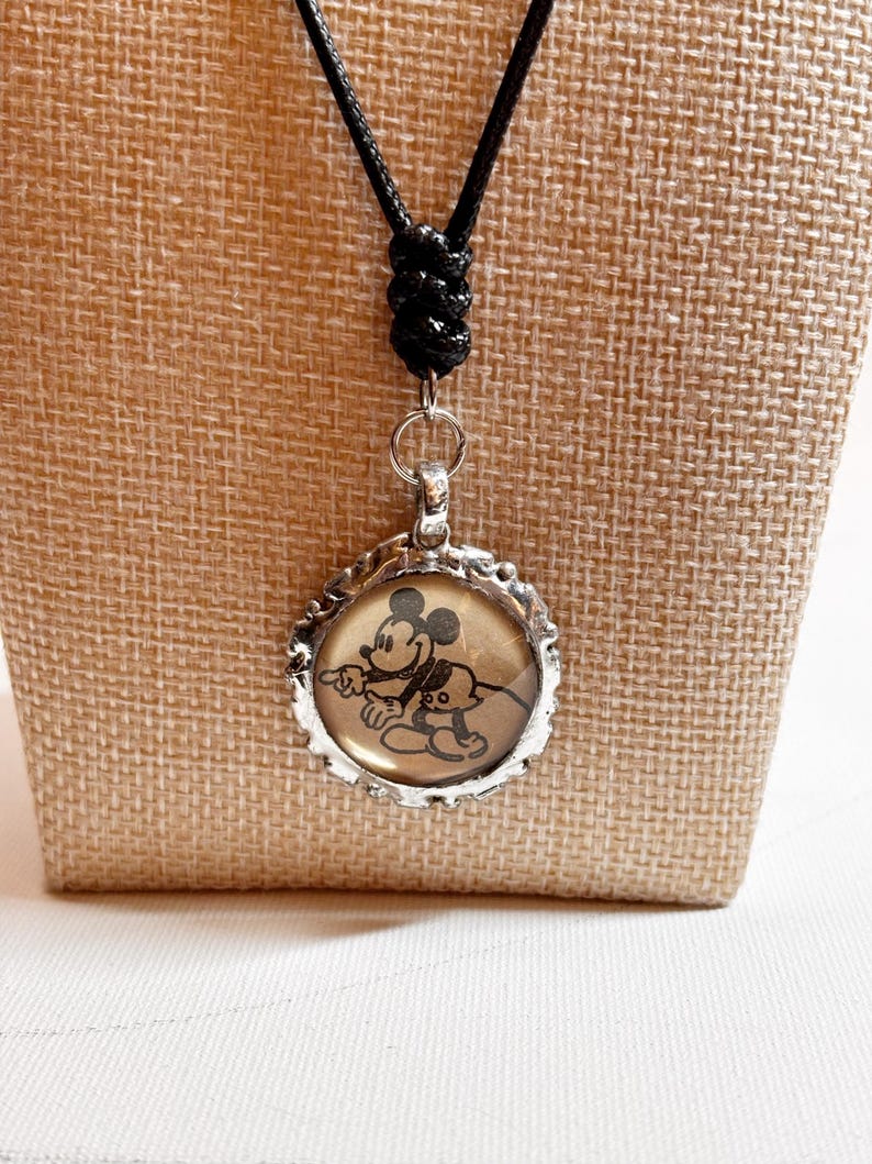 May include: A silver-toned pendant necklace featuring a circular charm with a black and white image of Mickey Mouse and Pluto. The charm is set in a decorative frame and hangs from a black cord necklace.