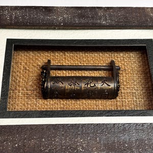 May include: A dark bronze-toned metal cylindrical puzzle lock featuring engraved Chinese characters. The lock has a horizontal bar across the top and is mounted within a rectangular frame with a textured, tan-colored fabric backing.