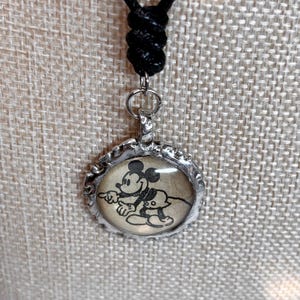 May include: A black cord necklace with a silver-colored pendant. The pendant features a black and white image of Mickey Mouse and Pluto. The pendant is round with a decorative edge.