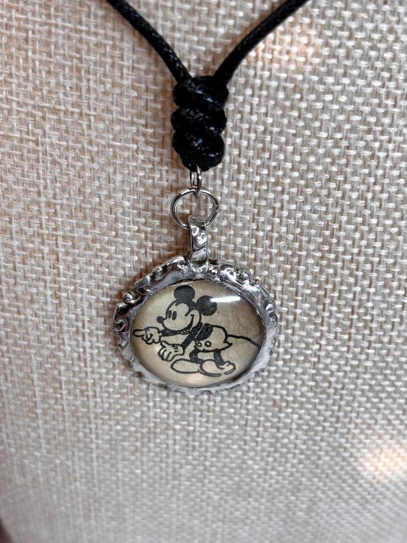 May include: A black cord necklace with a silver-colored pendant. The pendant features a black and white image of Mickey Mouse and Pluto. The pendant is round with a decorative edge.