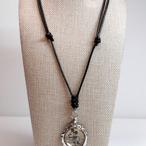 May include: A black cord necklace featuring a round pendant with a vintage Mickey Mouse image. The pendant has a silver-colored, scalloped edge. The necklace is displayed on a neutral-toned mannequin.
