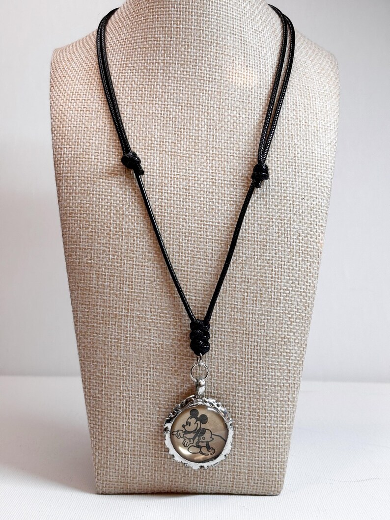 May include: A black cord necklace featuring a round pendant with a vintage Mickey Mouse image. The pendant has a silver-colored, scalloped edge. The necklace is displayed on a neutral-toned mannequin.
