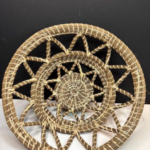 Pode incluir: Uma cesta redonda tran&ccedil;ada com um centro em espiral e um padr&atilde;o de treli&ccedil;a aberto em forma de estrela nas laterais. A cesta &eacute; feita de fibras naturais castanhas com fio de liga&ccedil;&atilde;o de cor creme mais claro por toda a estrutura.