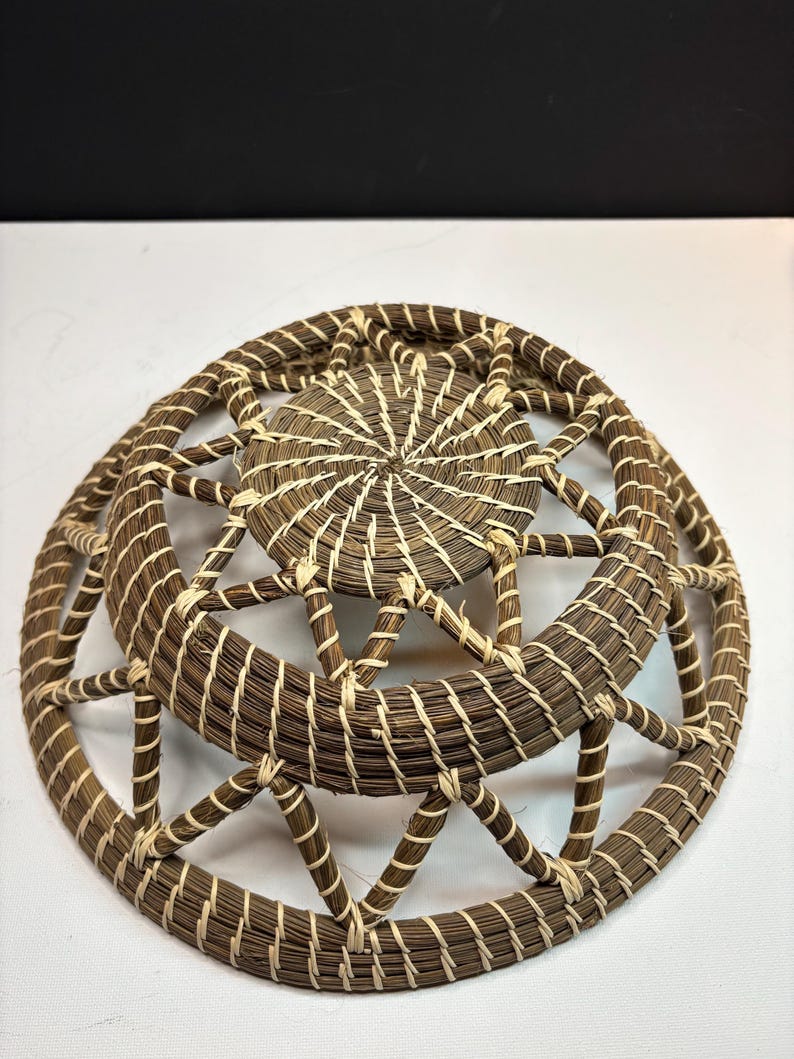Pode incluir: Uma cesta redonda tran&ccedil;ada feita de um material fibroso castanho-escuro, enrolado e unido com fio de cor creme claro. A estrutura inclui uma base circular s&oacute;lida e um padr&atilde;o de treli&ccedil;a geom&eacute;trica aberto em dire&ccedil;&atilde;o &agrave; borda.