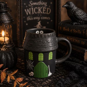 May include: A black ceramic mug shaped like a haunted house, featuring a green door, three green windows, a white spiderweb, two white skulls, and a white broom. The rim of the mug has a textured roof design with two white eyes peeking out.