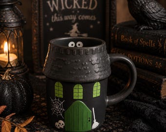 Pode incluir: Uma caneca de cerâmica preta em forma de casa assombrada, com uma porta verde, três janelas verdes, uma teia de aranha branca, dois crânios brancos e uma vassoura branca. A borda tem um design de telhado texturizado com dois olhos.