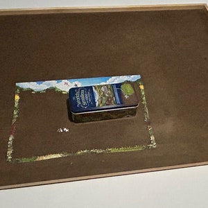 May include: A brown wooden jigsaw puzzle board with a raised edge. A partially completed jigsaw puzzle is on the board. A blue and white tin with the text "Wonderland" is on top of the puzzle.