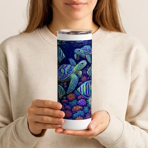 May include: A white and stainless steel insulated tumbler featuring a vibrant underwater scene. The design includes sea turtles, colorful fish, and coral reefs in shades of blue, green, and purple. The tumbler is held by a person.