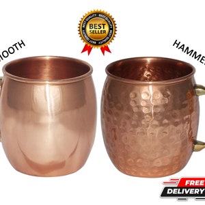 以下が含まれることがあります： 銅製のモスコミュールマグカップ2つ。1つは滑らかで、もう1つはハンマー加工が施されています。真鍮製のハンドルが付いています。マグカップは並んで表示され、テクスチャの違いを示しています。画像には「Smooth」と「Hammered」というテキストが含まれています。