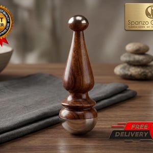 Puede incluir: Una herramienta de masaje de madera marrón oscuro con una base plateada. La herramienta tiene una parte superior redondeada y una forma cónica. La imagen también incluye un paño gris, una botella pequeña y una insignia de "Best Seller".