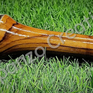 May include: A wooden pestle with a polished, metallic head and a rounded handle, resting on a bed of green grass. The wood grain is visible, and the pestle is designed for grinding spices or herbs. The text "Spornzo Creation" is visible.