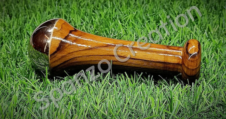 May include: A wooden pestle with a polished, metallic head and a rounded handle, resting on a bed of green grass. The wood grain is visible, and the pestle is designed for grinding spices or herbs. The text "Spornzo Creation" is visible.