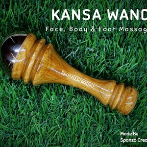 May include: A Kansa wand face, body, and foot massager with a wooden handle and a metal ball. The text "KANSA WAND" and "Face, Body & Foot Massager" are visible. The wand is on a green grassy surface.