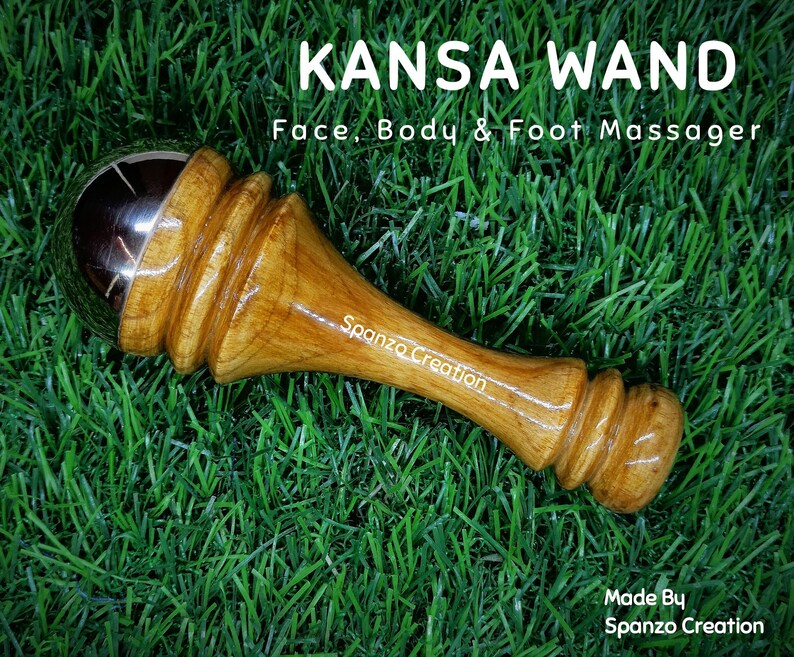 May include: A Kansa wand face, body, and foot massager with a wooden handle and a metal ball. The text "KANSA WAND" and "Face, Body & Foot Massager" are visible. The wand is on a green grassy surface.