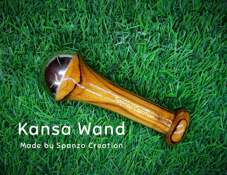 May include: A Kansa wand with a polished metal sphere and a wooden handle. The handle is made of light brown wood with dark brown grain patterns. The text "Kansa Wand" and "Made by Spanzo Creation" are visible.
