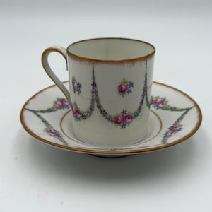 May include: A white teacup and saucer set with a gold rim. The cup and saucer are decorated with a floral garland design in pink, blue, and green. The cup has a curved handle.